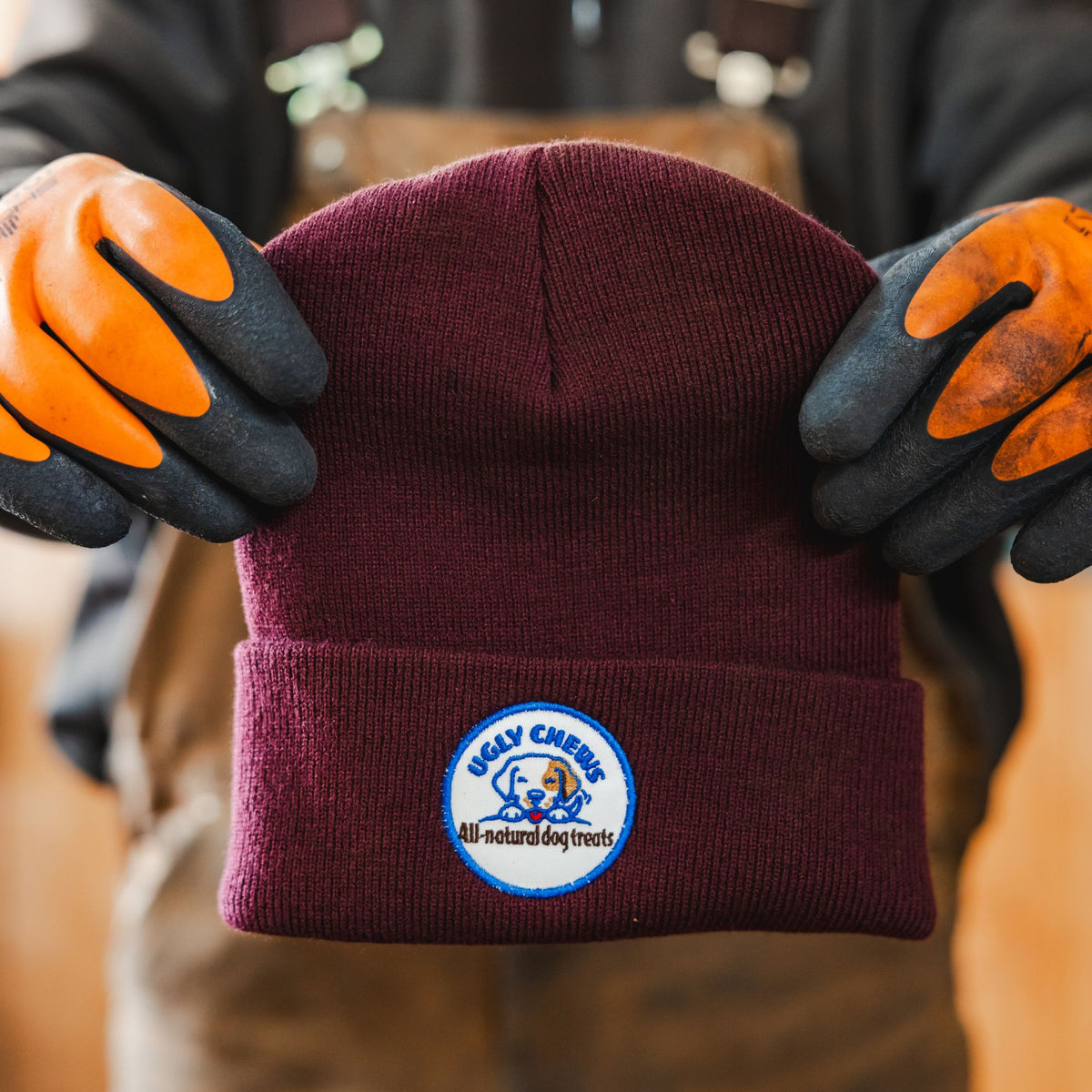 Gloved hands holding up a maroon Ugly Chews beanie